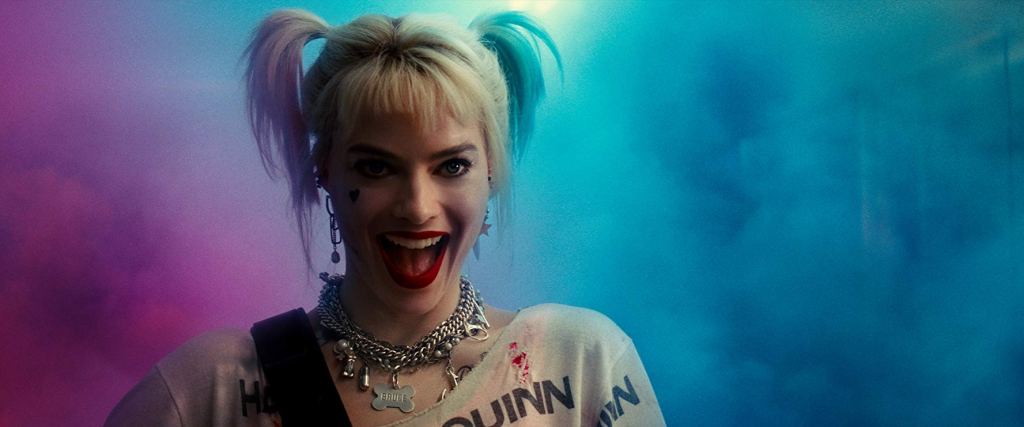 Photograph of Margot Robbie as Harley Quinn.  She has light skin, short blond hair in pigtails, mismatched dangle earring, close fitting metal charm necklace, and a blood splattered white t-shirt.  Her mouth is open in a wide grin or laugh.  